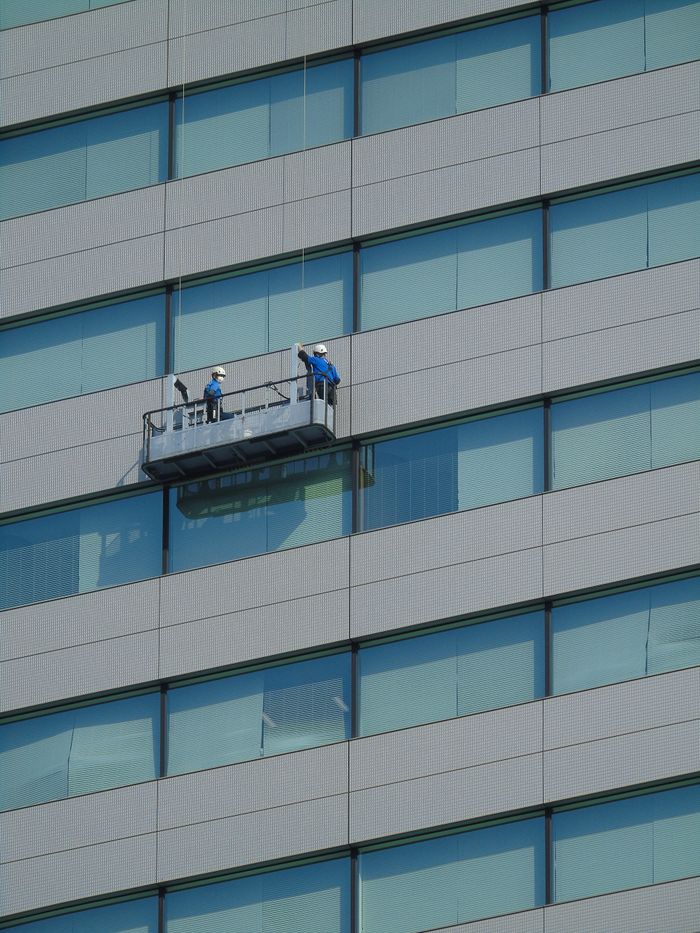 about-01 Two workers are cleaning windows on the facade of a modern glass building from a suspended platform.